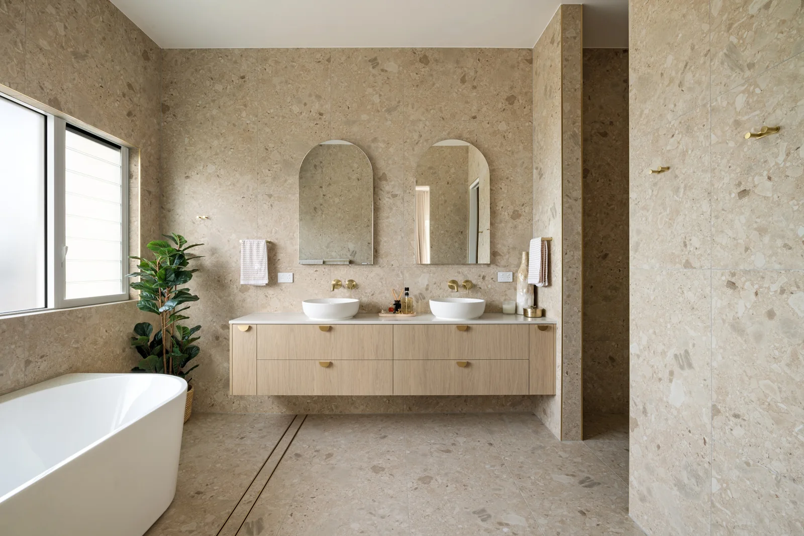 Warm stone bathroom with twin basins, arched mirrors and a freestanding bath